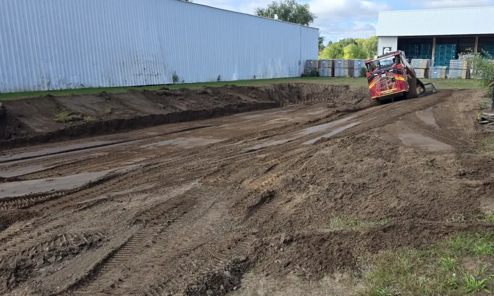 Construction equipment grading soil for a new retention pond beside a large metal warehouse building.