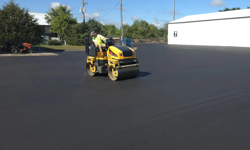 Worker operating a double drum roller to compact freshly laid asphalt on a newly paved parking lot